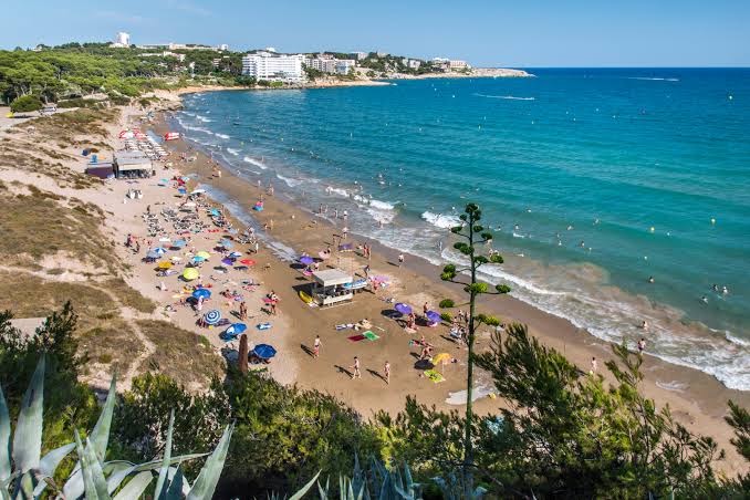 Salovas smilšu pludmale netālu no 4R Playa Park viesnīcas — tirkīzzils ūdens, atpūtnieki un piekrastes veģetācija saulainā dienā.