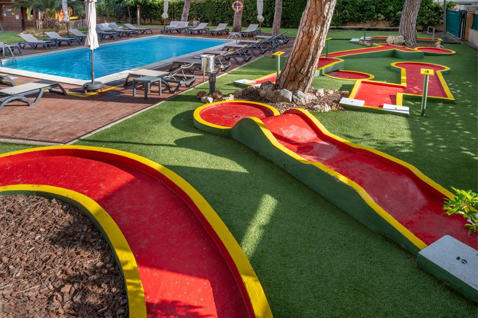 4R Playa Park āra baseins un minigolfa laukums ar sarkanām trasēm zaļā zālājā, ko ieskauj priedes un sauļošanās krēsli