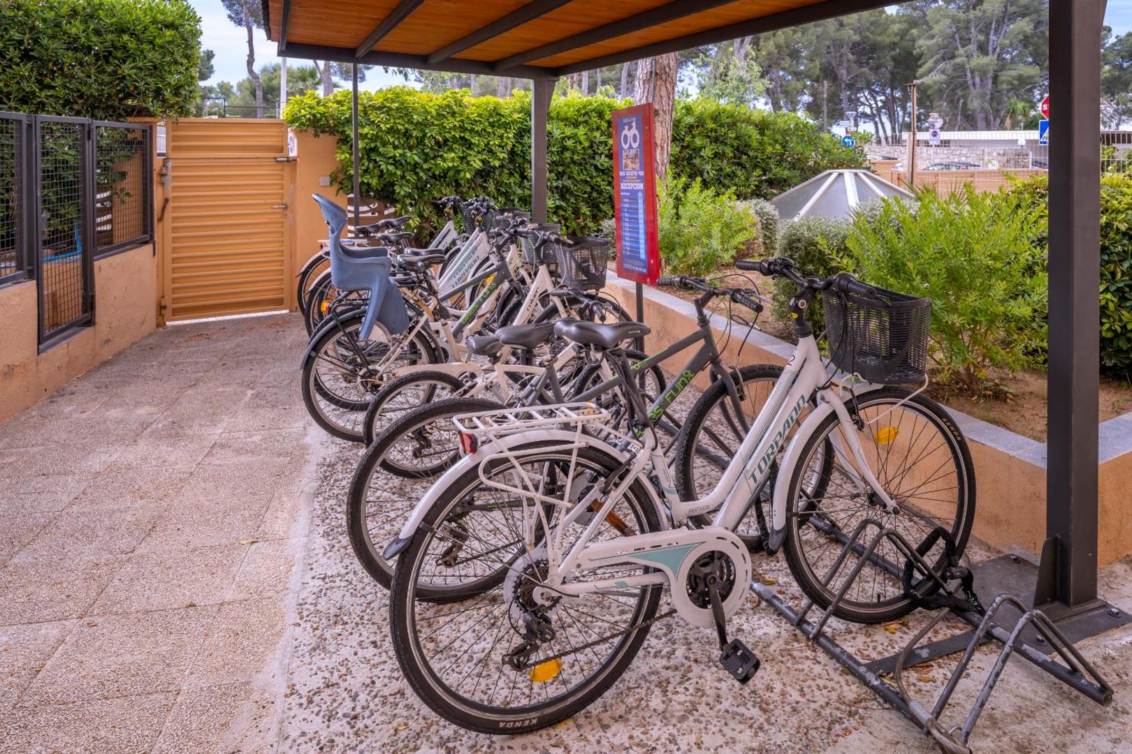 Velosipēdu noma viesnīcā 4R Playa Park — dažāda veida velosipēdi ar bērnu sēdeklīšiem novietoti zem nojumes aktīvai atpūtai