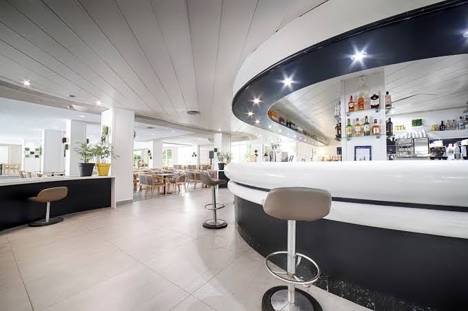 Ohtels Belvedere modernais lobby bārs ar pusapaļu baltu leti, augstiem bāra krēsliem un plašu dzērienu izvēli fonā