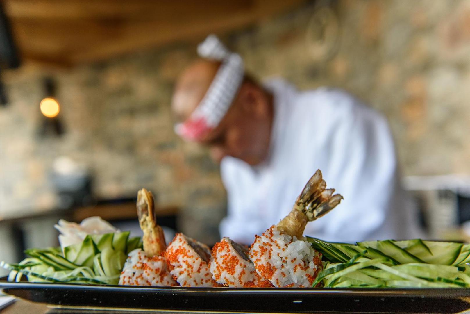 Royal Marmin Bay restorāna šefpavārs gatavo suši — tuvplāns ar tempura garnelēm un ikriem uz akmens sienas fona