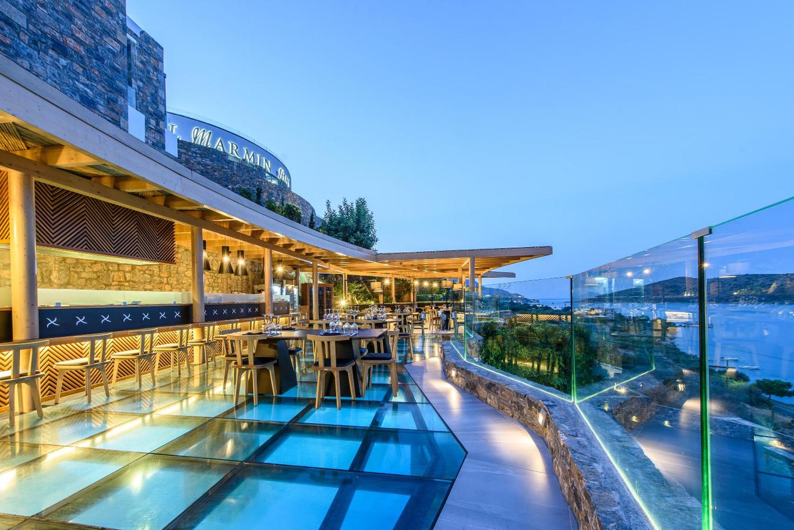 Royal Marmin Bay Boutique & Art Hotel restorāns ar stikla grīdu un modernu dizainu, no kura paveras skats uz jūru saulrietā.