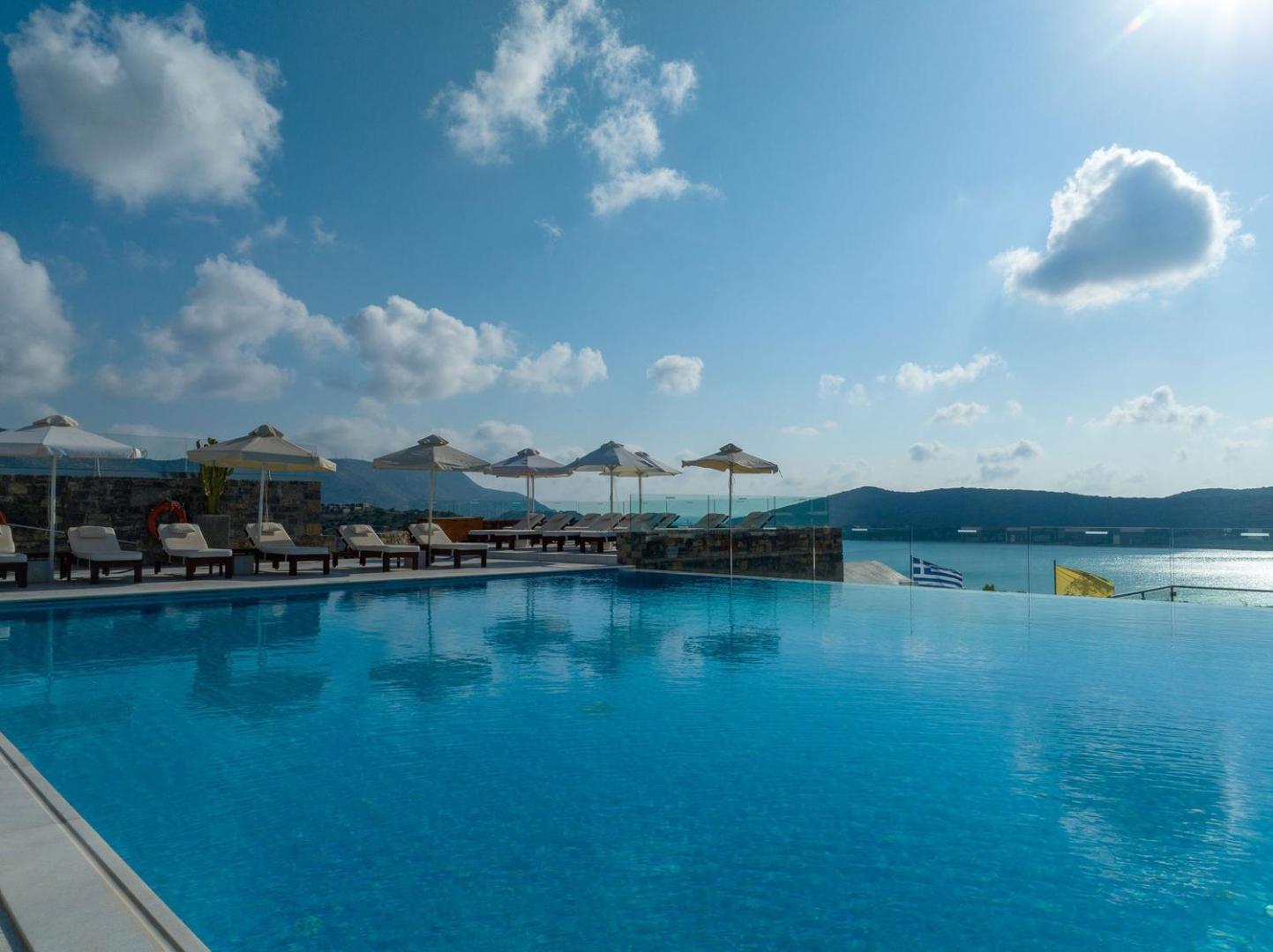 Royal Marmin Bay Boutique & Art Hotel bezgalības baseina ūdens virsma ar saules atspulgiem un saulessargiem piekrastes fonā.
