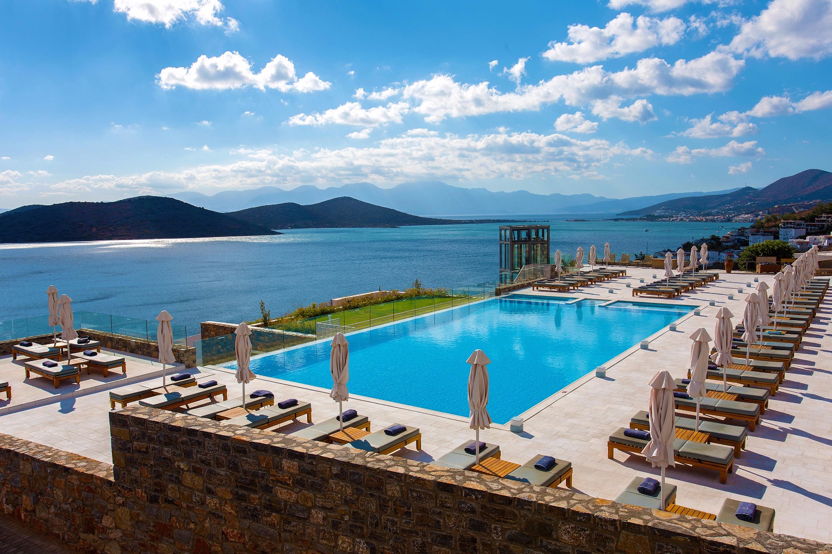 Royal Marmin Bay Boutique & Art Hotel galvenais āra baseins ar sauļošanās krēsliem, saulessargiem un panorāmas skatu uz Krētas piekrasti.
