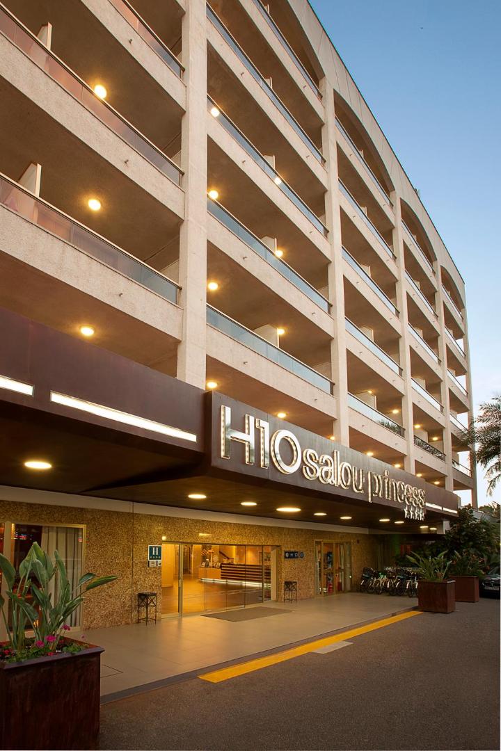 H10 Salou Princess viesnīcas ieeja un fasāde vakarā Salovā, Spānijā — apgaismota izkārtne un balkonu rindas piekrastes kūrortā