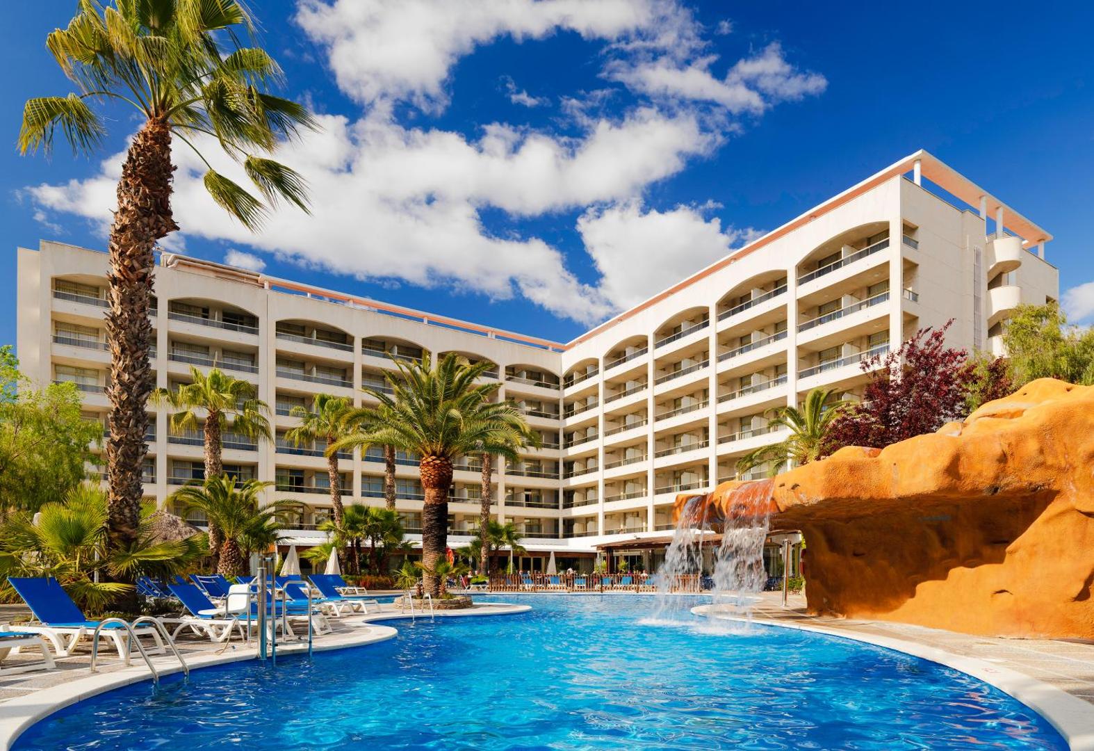 H10 Salou Princess āra baseins ar mākslīgo ūdenskritumu, sauļošanās krēsliem un palmām pie viesnīcas galvenās ēkas Salovā