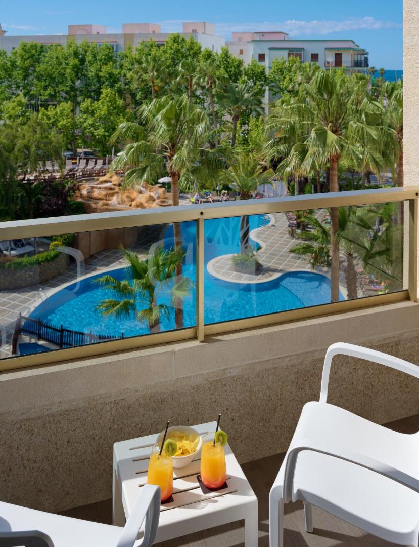 Skats no H10 Salou Princess balkona uz zilo āra baseinu, palmām un kūrorta teritoriju saulainā dienā
