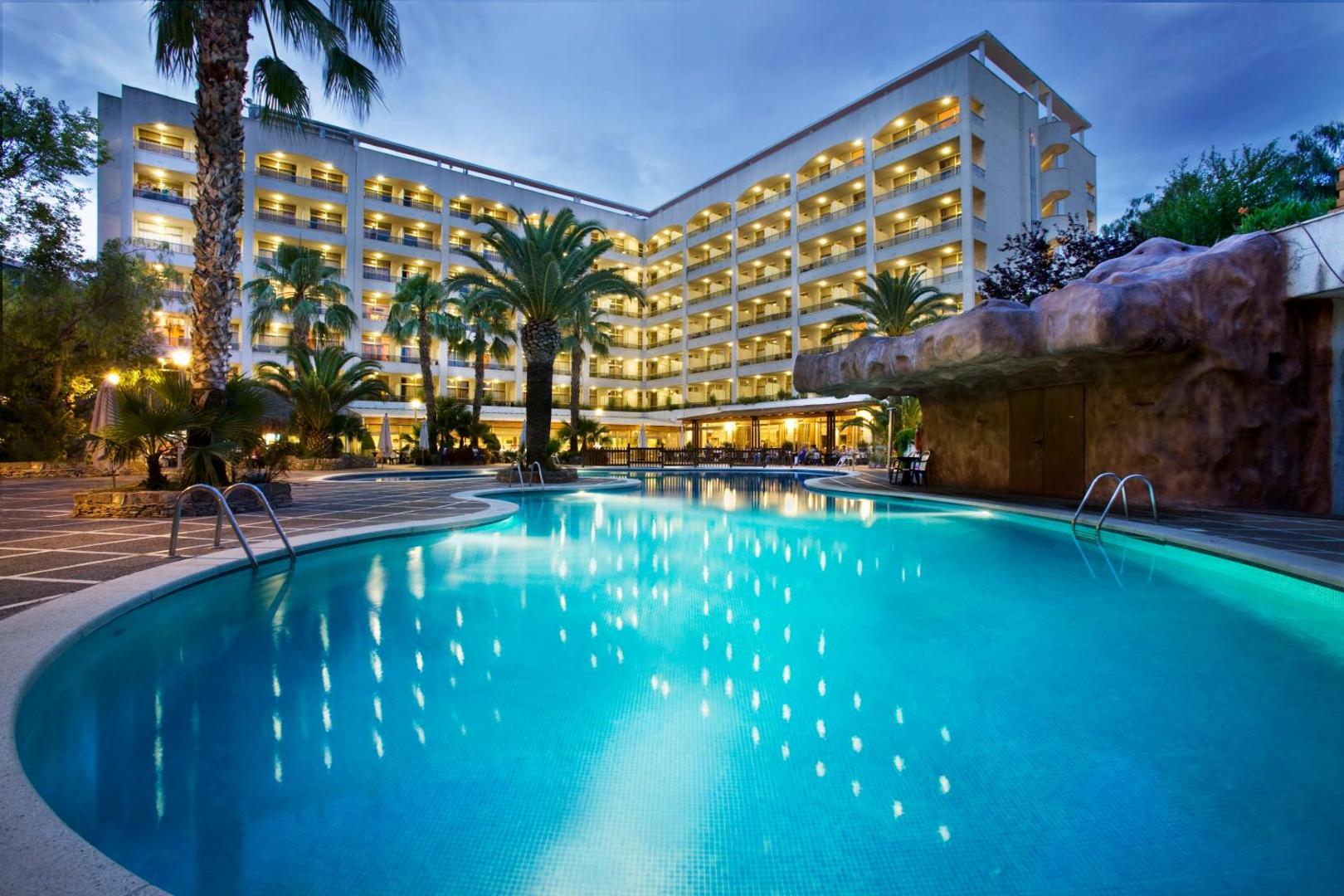 H10 Salou Princess viesnīcas ēka un izgaismots āra baseins vakarā ar palmām un dekoratīvām klintīm Kostadoradas piekrastē