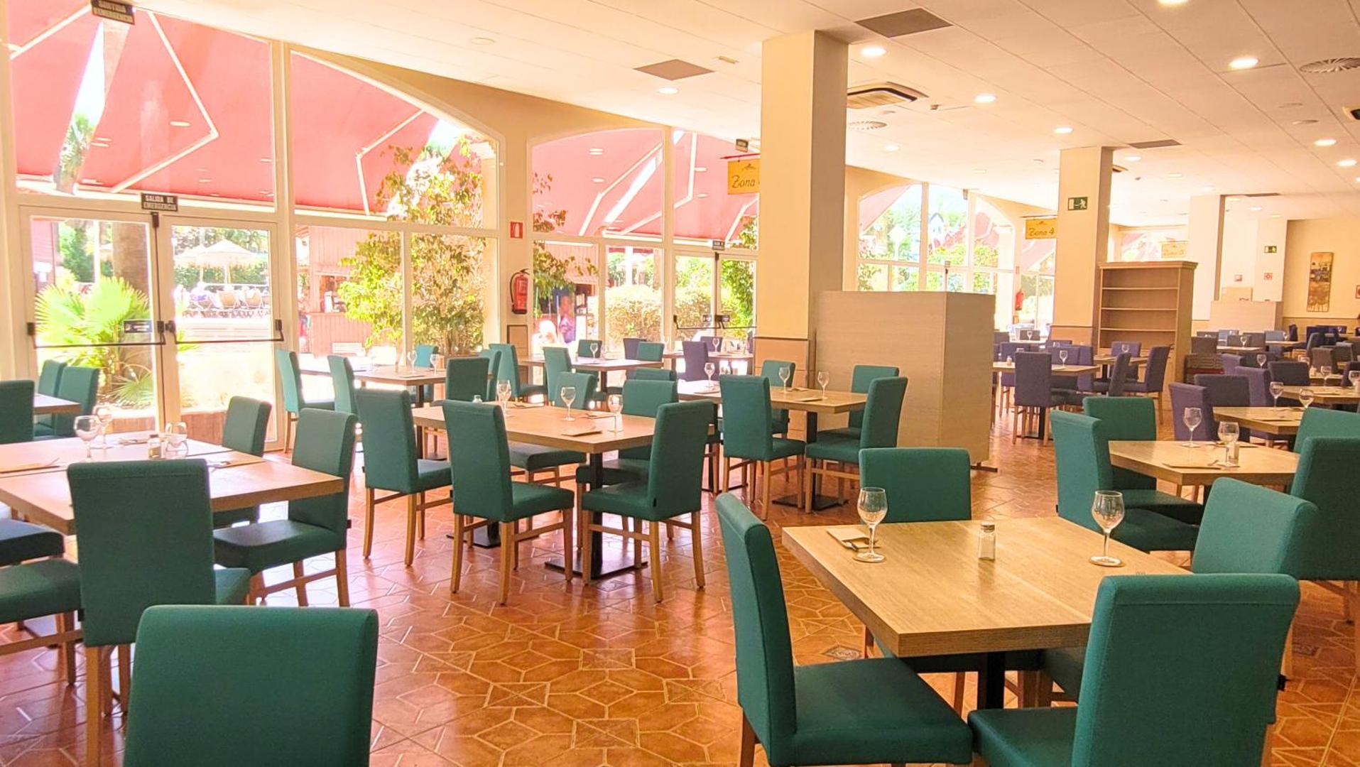Ohtels La Hacienda plašā ēdamzāle ar lieliem logiem, tirkīza krāsas krēsliem un skatu uz saulaino āra terasi.