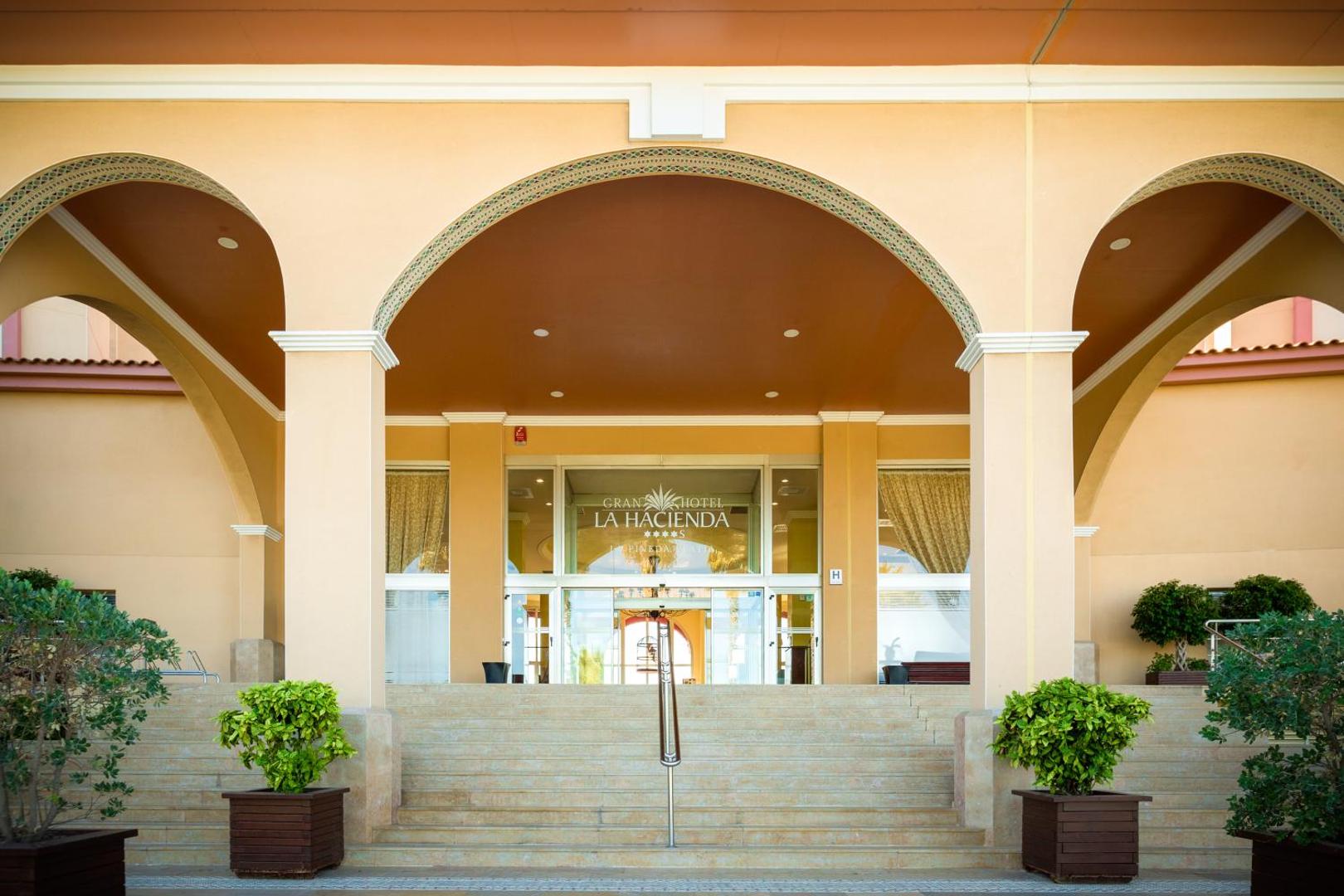 Tuvplāns uz Ohtels La Hacienda ieeju ar monumentālām arkām un kāpnēm, kas ved uz viesnīcas vestibilu