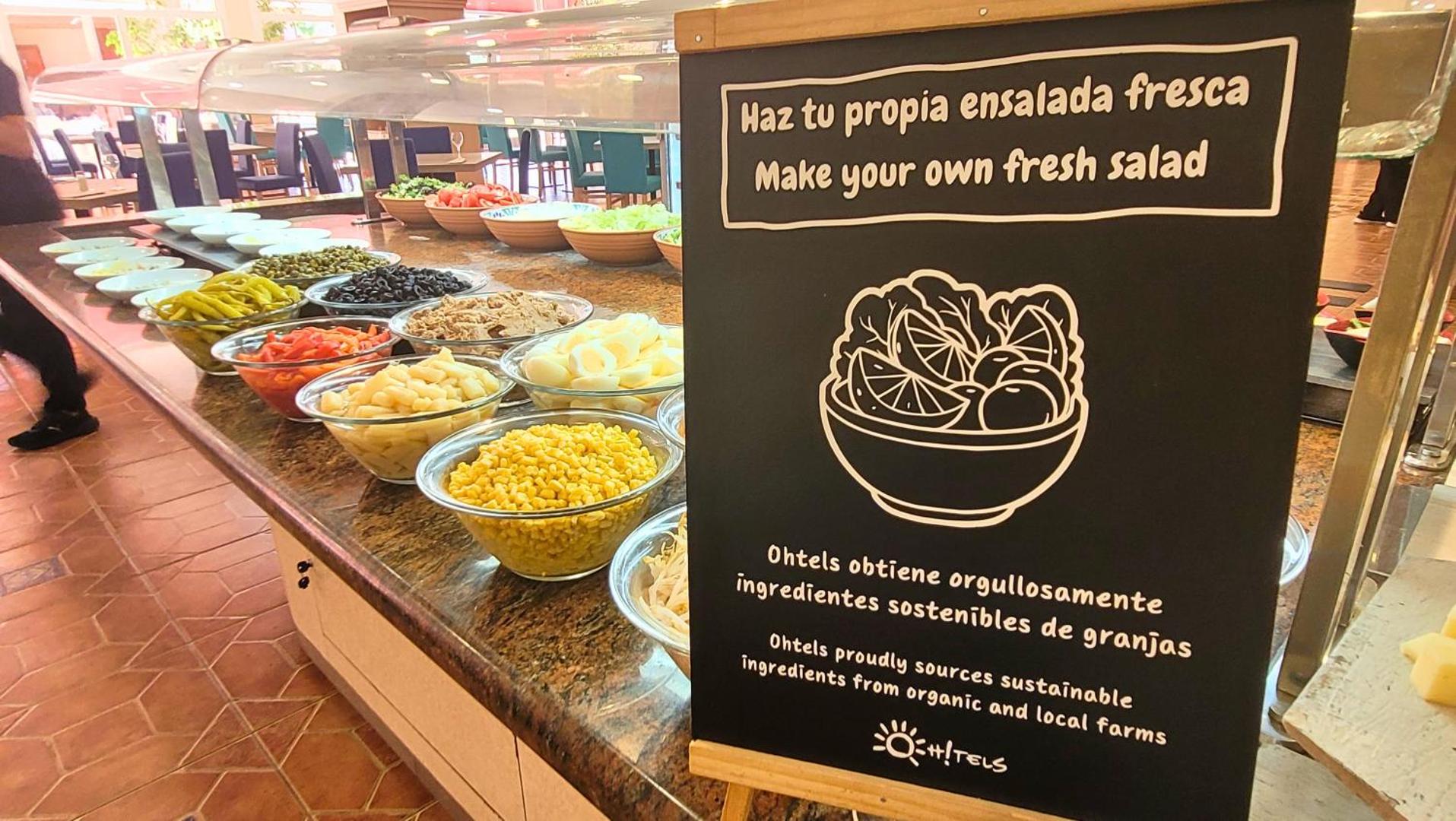 Ohtels La Hacienda bufetes restorāna salātu bārs ar svaigām sastāvdaļām un informatīvu zīmi par vietējiem produktiem