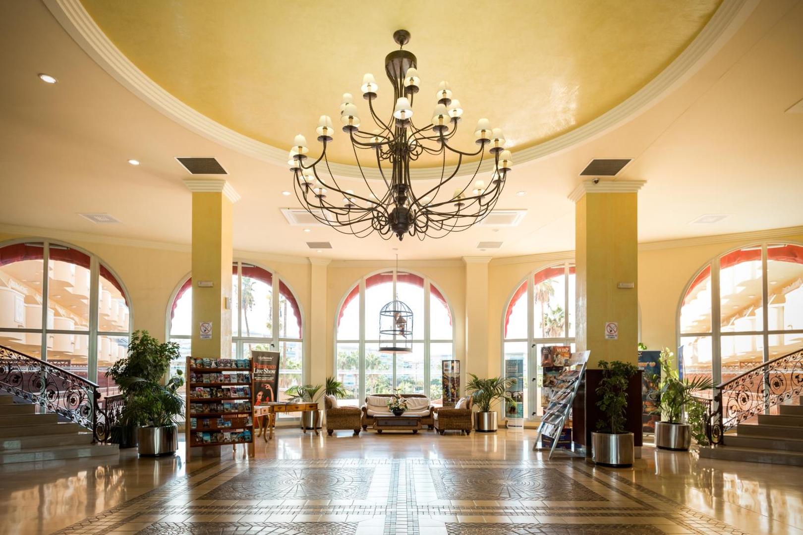 Ohtels La Hacienda viesnīcas lobby ar augstiem griestiem, greznu lustru, lieliem arkveida logiem un atpūtas zonu ar dīvāniem.