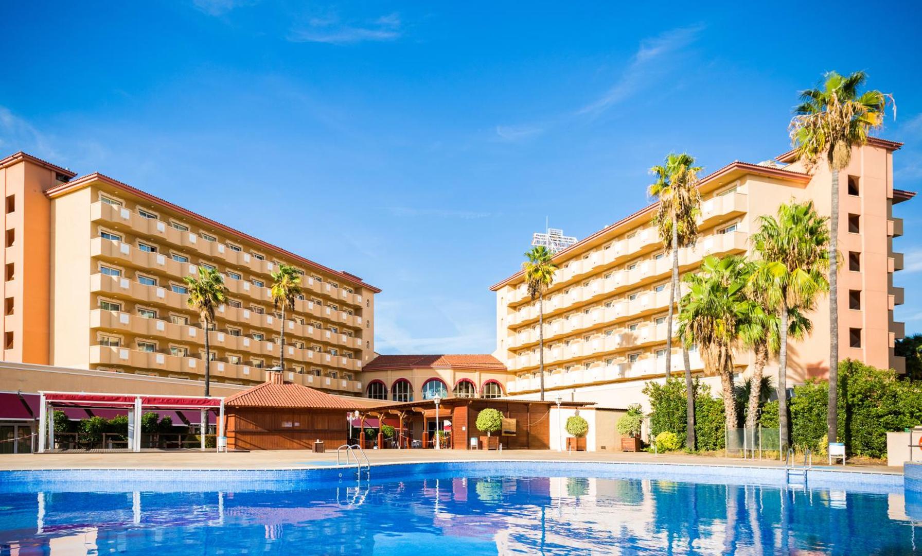Ohtels La Hacienda viesnīcas ēkas un lielais āra baseins saulainā dienā, ko ieskauj palmas un plaša atpūtas zona.