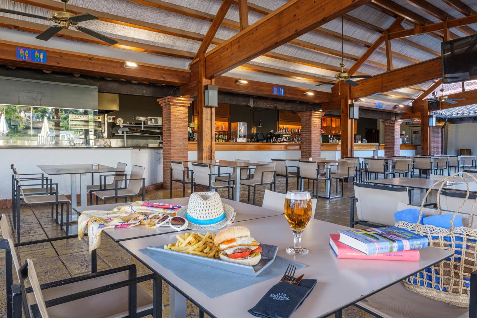 Estival Eldorado Resort restorāna terase ar koka sijām — galds ar burgeri, frī kartupeļiem un atspirdzinošu dzērienu mājīgā atmosfērā