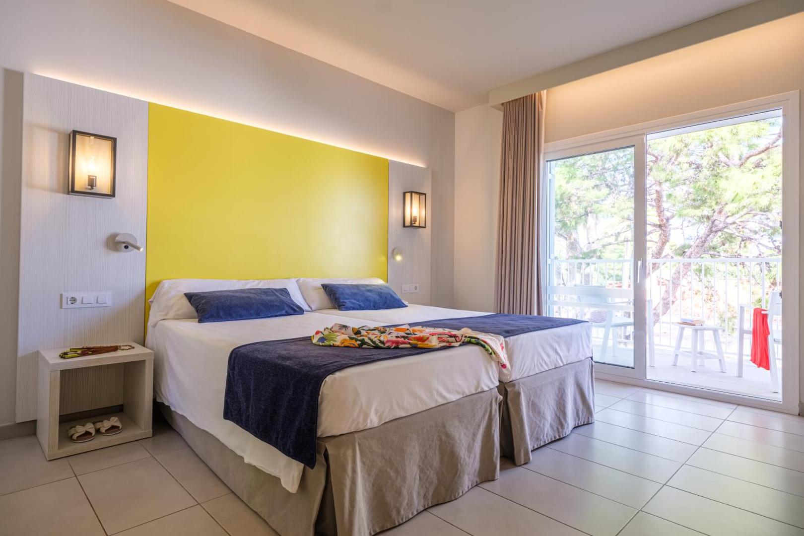Estival Eldorado Resort numurs ar dzeltenu sienas akcentu, divām gultām un plašām stikla durvīm uz balkonu priežu ielokā