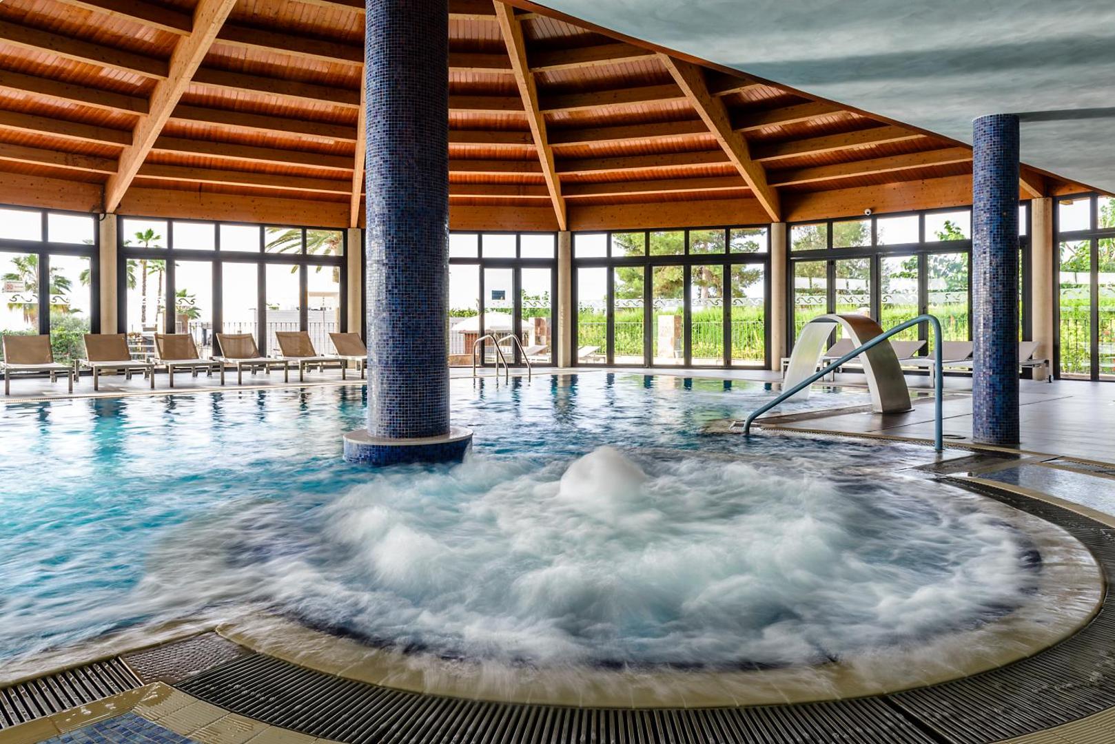 Estival Eldorado Resort iekštelpu spa baseins ar hidromasāžas zonu un koka griestu konstrukciju Kambrilsā, Spānijā — atpūtas krēsli pie lieliem logiem.