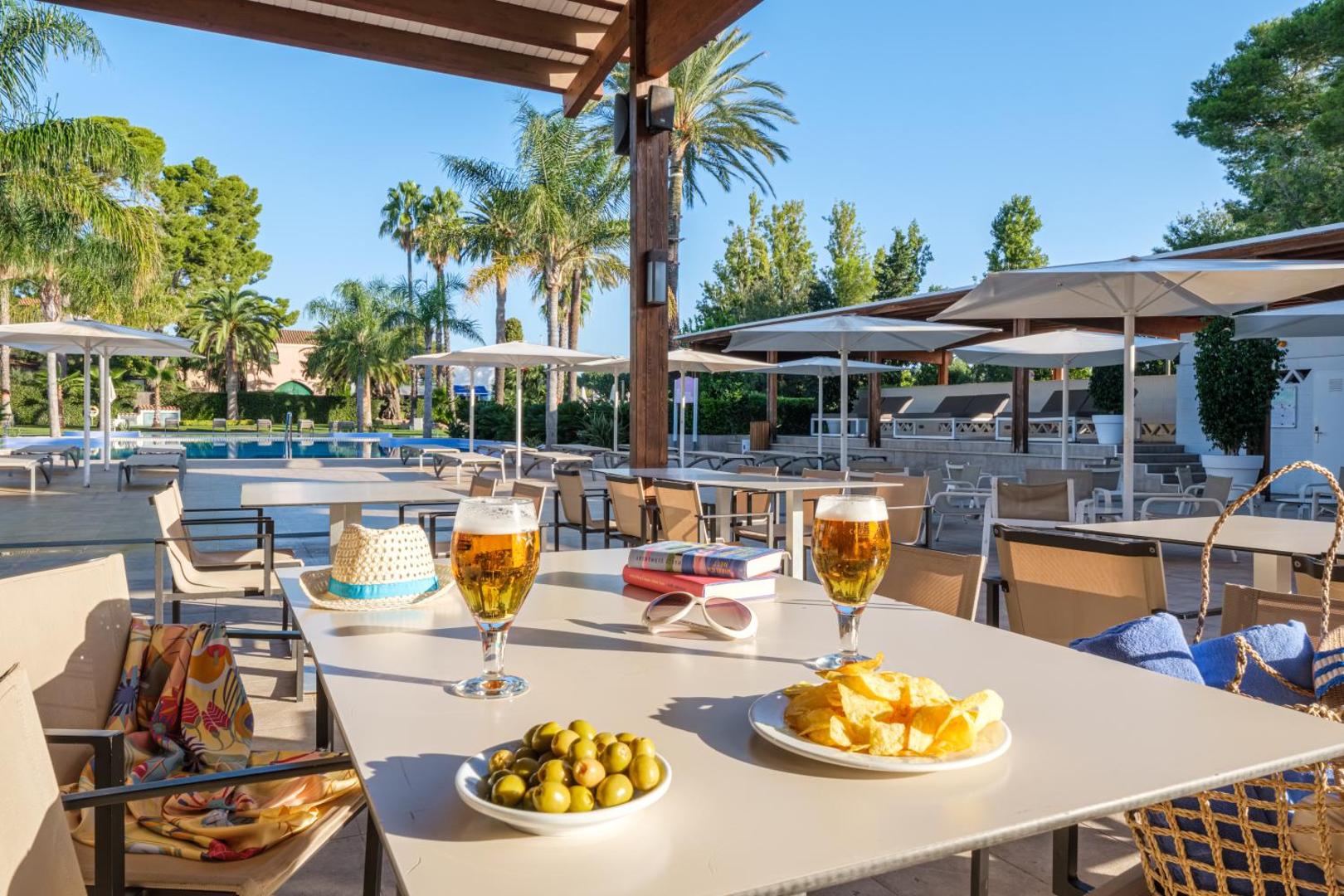 Estival Eldorado Resort āra terase ar skatu uz baseinu un palmām — uz galda olīvas un dzērieni saulainā pēcpusdienā