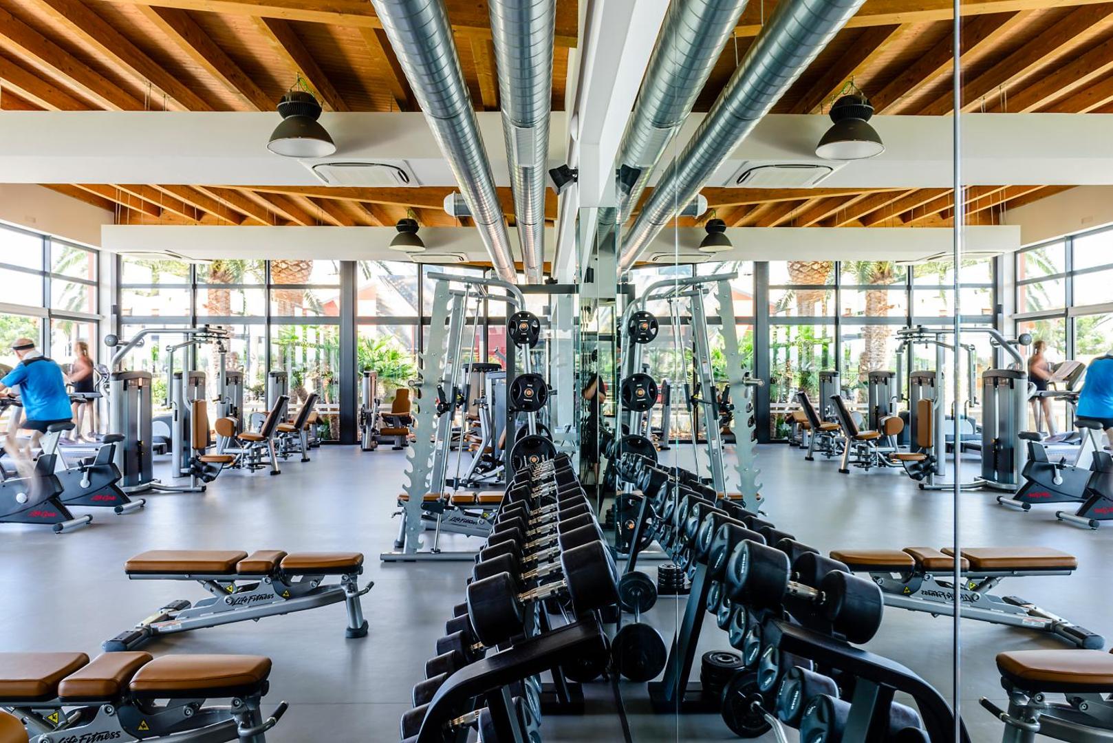 Estival Eldorado Resort moderns fitnesa centrs ar Life Fitness spēka trenažieriem, hantelēm un panorāmas logiem uz dārzu Kambrilsā, Spānijā.