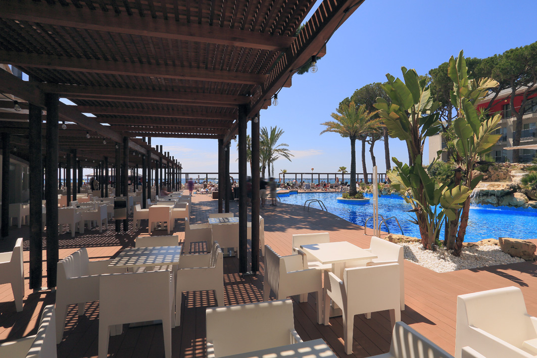Estival Centurion Playa viesnīcas āra baseins ar koka terasi un restorāna zonu Kambrilsā, Spānijā — palmas un sauļošanās krēsli pie ūdens