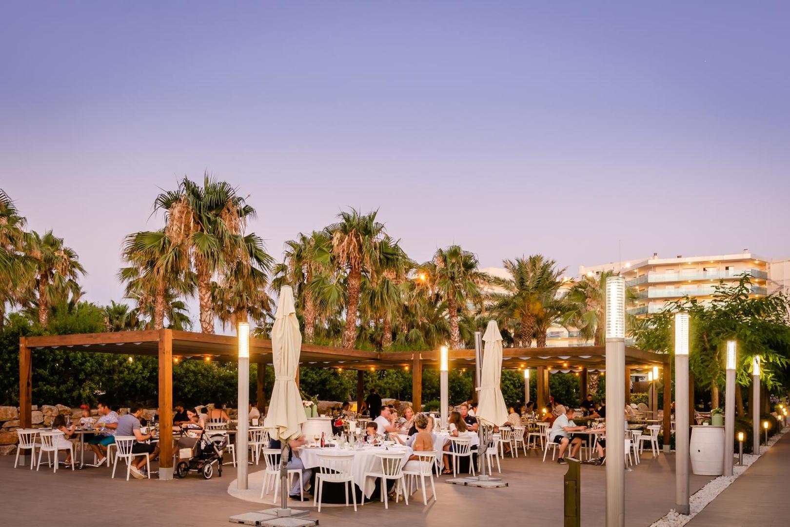 Viesnīcas Gran Palas āra terases restorāns vakara apgaismojumā ar palmām un balti klātiem galdiem kūrorta teritorijā.