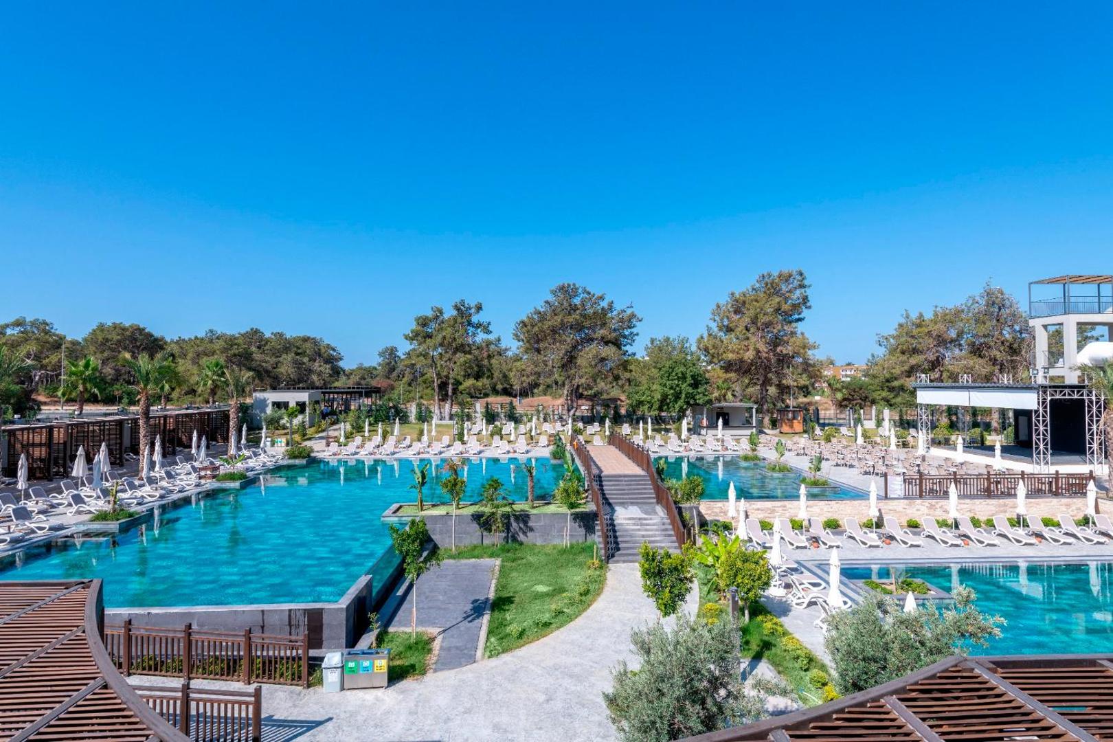 Sorgun Akadia Hotel Luxury dārza zona ar baseiniem, koka tiltiņu un sauļošanās krēsliem — apkārtnē priedes un labiekārtoti celiņi