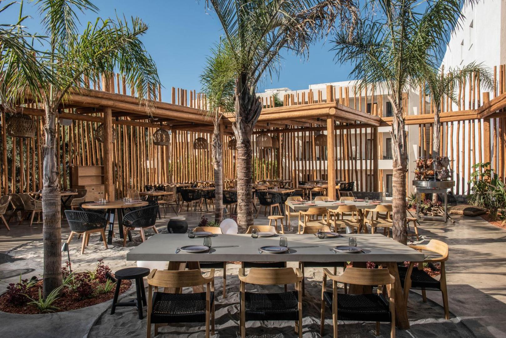 Indigo Inn Casa āra restorāna zona ar palmām un koka konstrukcijām Hersonisā — plaša terase ar smilšu segumu un dizaina krēsliem