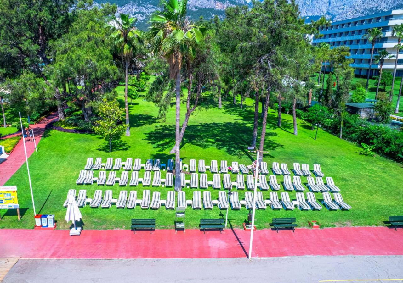 Kilikya Palace Goynuk viesnīcas dārzs ar daudziem sauļošanās krēsliem zālienā zem palmām un priedēm — skats no augšas