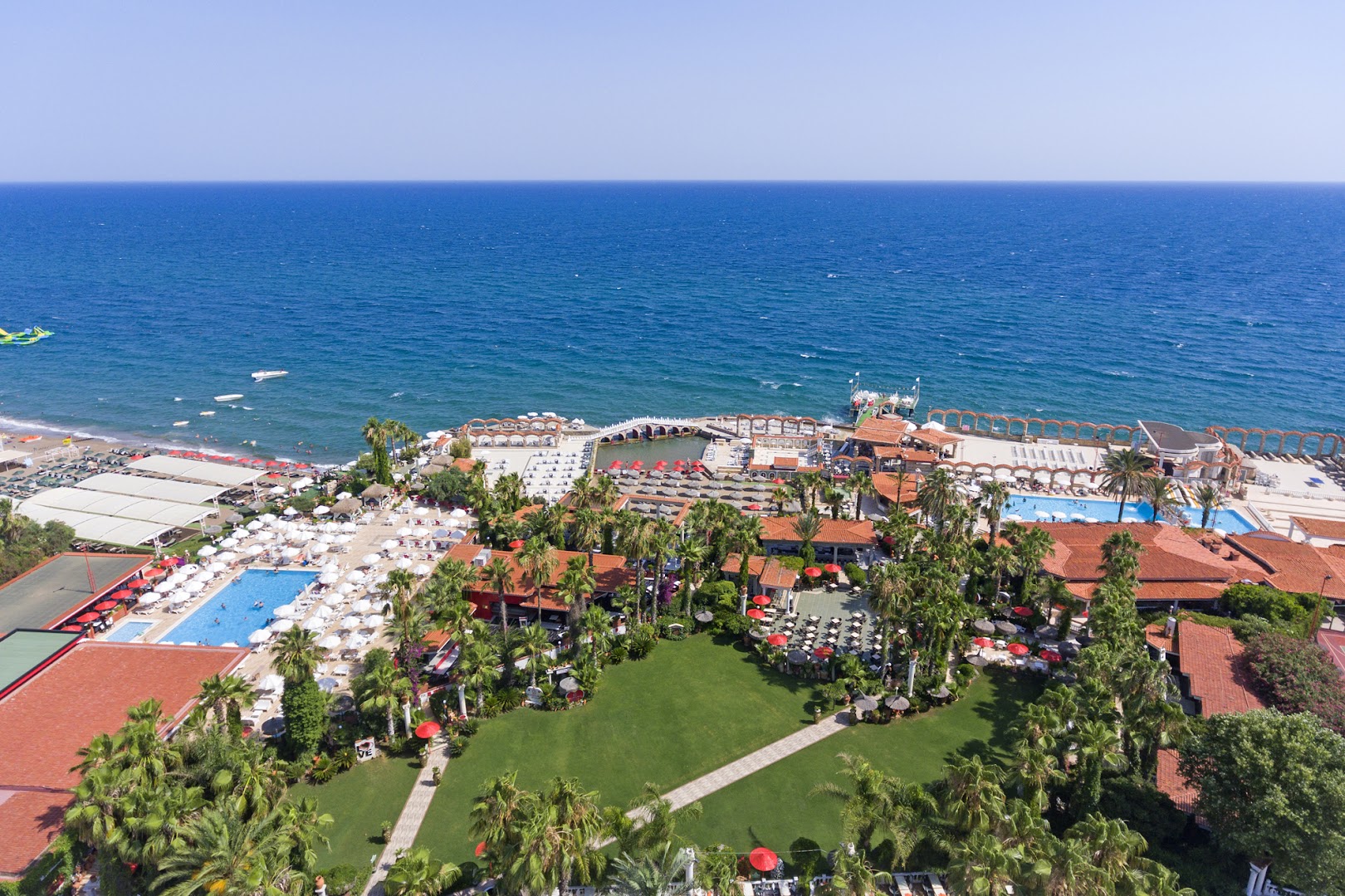 Club Hotel Sera dārza un baseinu zona no augšas — zaļš zālājs, palmas, vairāki atpūtas baseini un skats uz atklātu jūru