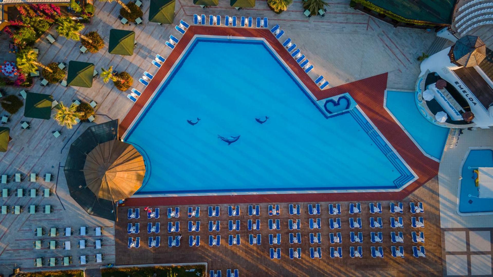 Emelda Sun Club galvenais baseins no putna lidojuma Kemeras reģionā — zils ūdens ar delfīnu mozaīkām un saulessargu zonu