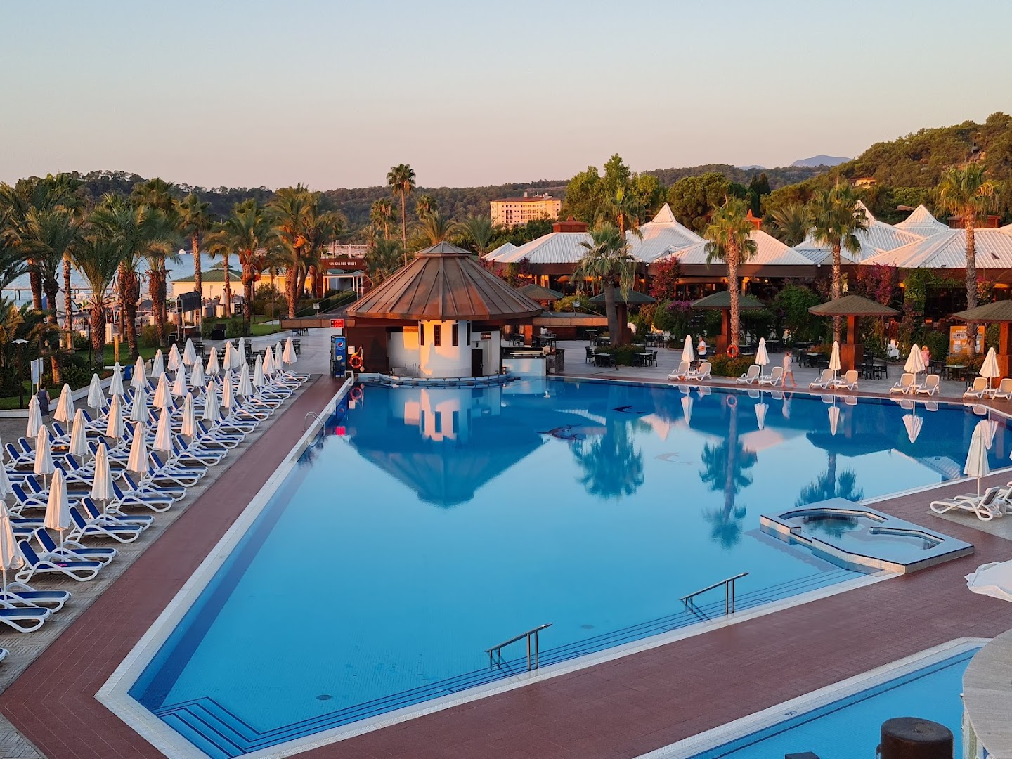 Emelda Sun Club āra baseins saulrietā ar skatu uz kalniem un bungalo stila ēkām Kemeras piekrastē, Turcijā