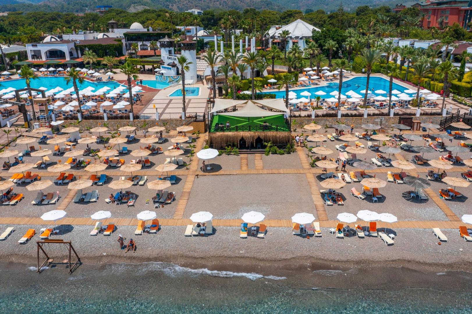 Emelda Sun Club viesnīcas teritorijas skats no augšas Kemerā, Turcijā — plaša pludmale ar saulessargiem un divi lieli āra baseini
