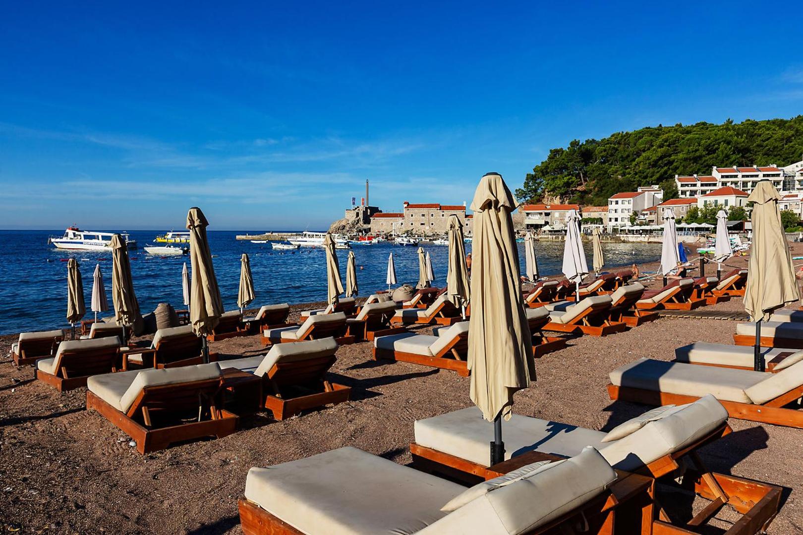 Sauļošanās krēsli un saulessargi Hotel Riva by Aycon pludmalē ar skatu uz Petrovacas līci un laivām zilajā jūrā