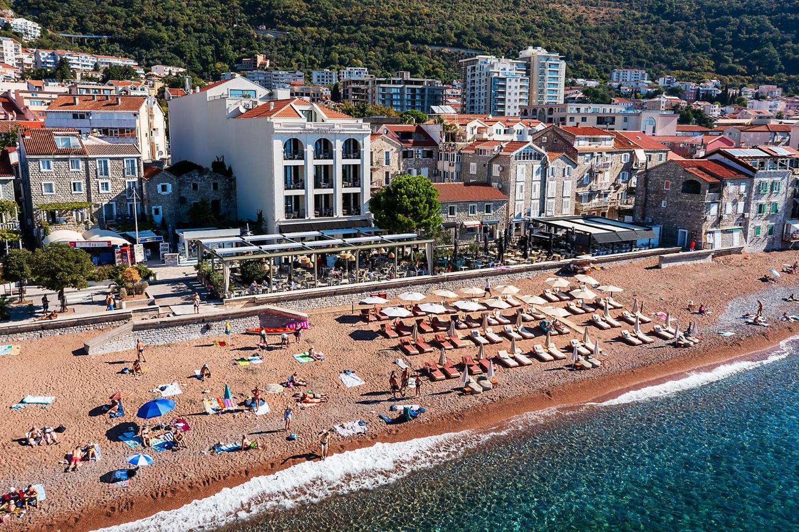 Aerofoto skats uz Riva by Aycon viesnīcu Petrovacā — plaša oļu pludmale ar atpūtas krēsliem, pilsētas apbūve un zaļi kalni.