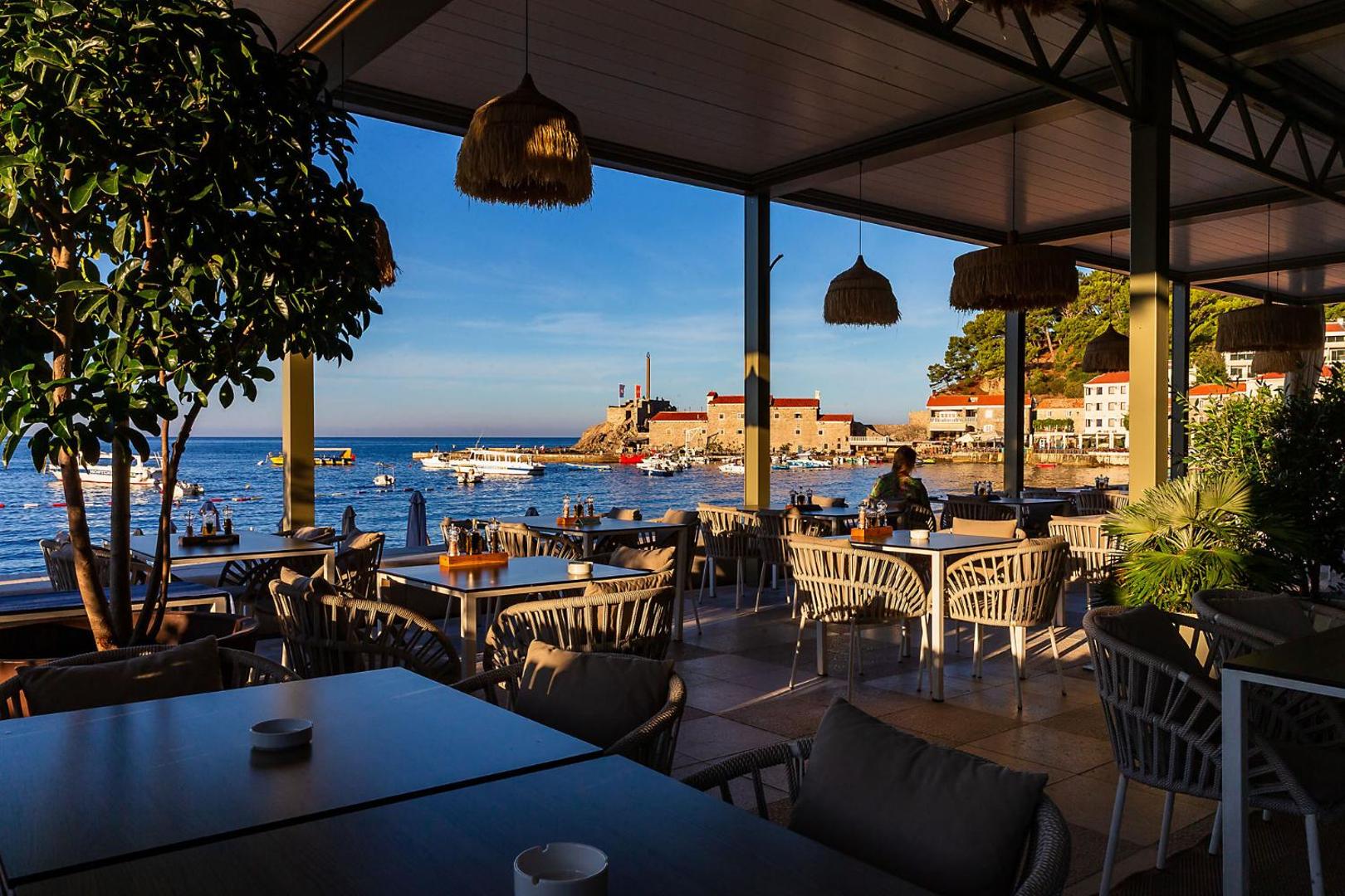 Skats no Hotel Riva by Aycon segtās terases uz Adrijas jūru un Petrovacas vēsturisko cietoksni saulrietā — stilīgi pīti krēsli