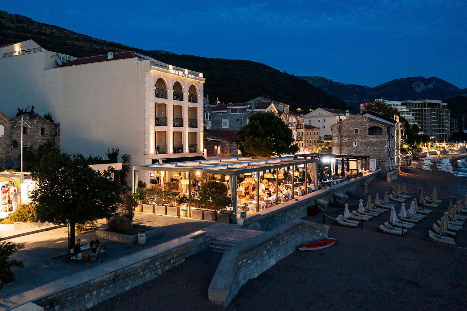 Riva by Aycon viesnīcas terase un restorāns vakarā — izgaismota āra zona ar galdiņiem pie pašas pludmales Petrovacā.