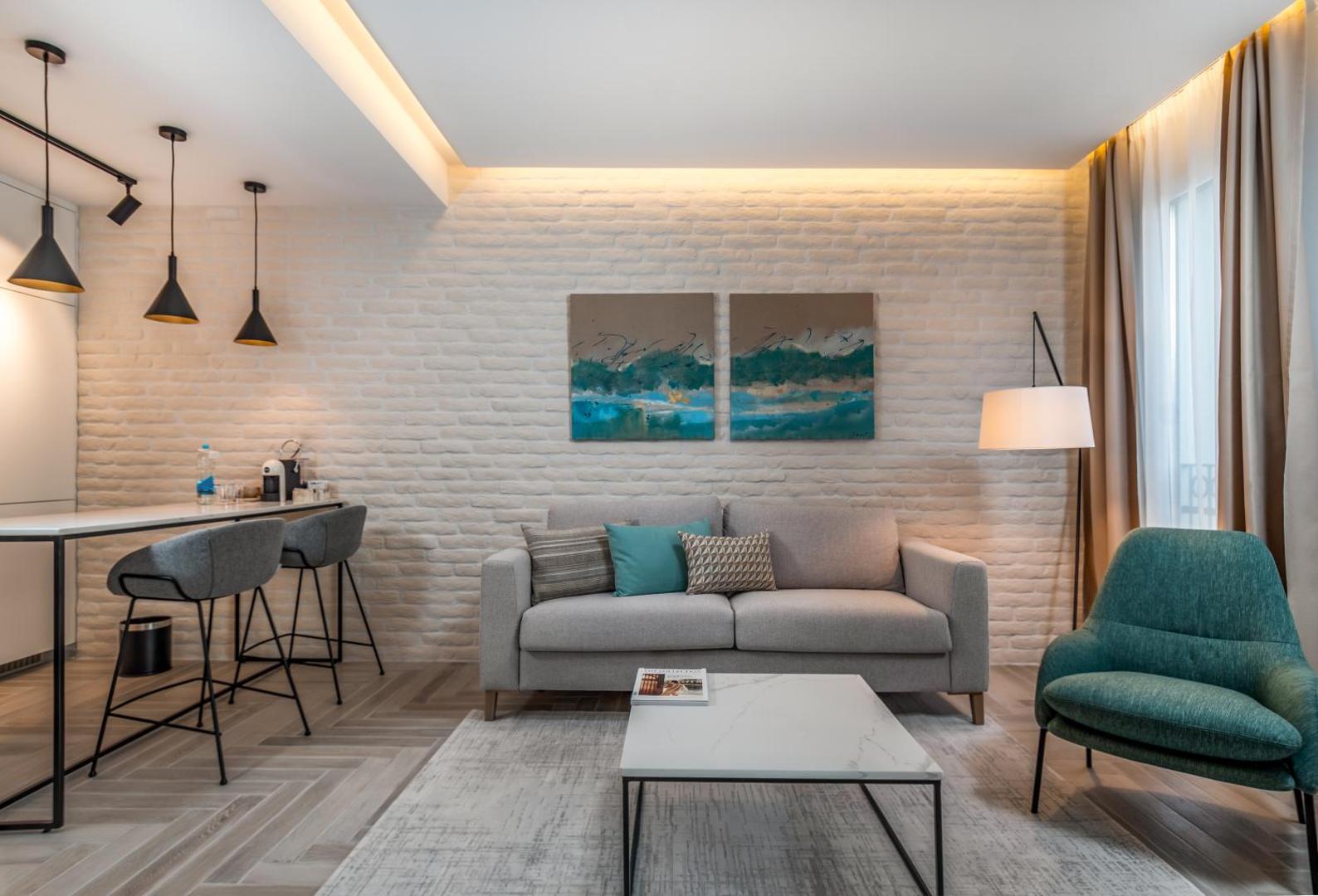 Lazure Hotel & Marina moderns viesnīcas numurs ar pelēku dīvānu, dekoratīviem spilveniem un nelielu virtuves zonu Melnkalnes piekrastē
