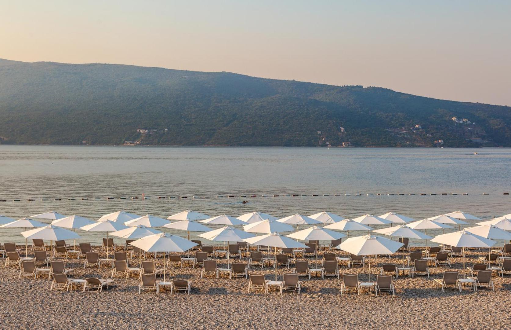 Lazure Hotel & Marina plašā oļu pludmale ar daudziem baltiem saulessargiem un krēsliem pie rāmiem Adrijas jūras ūdeņiem