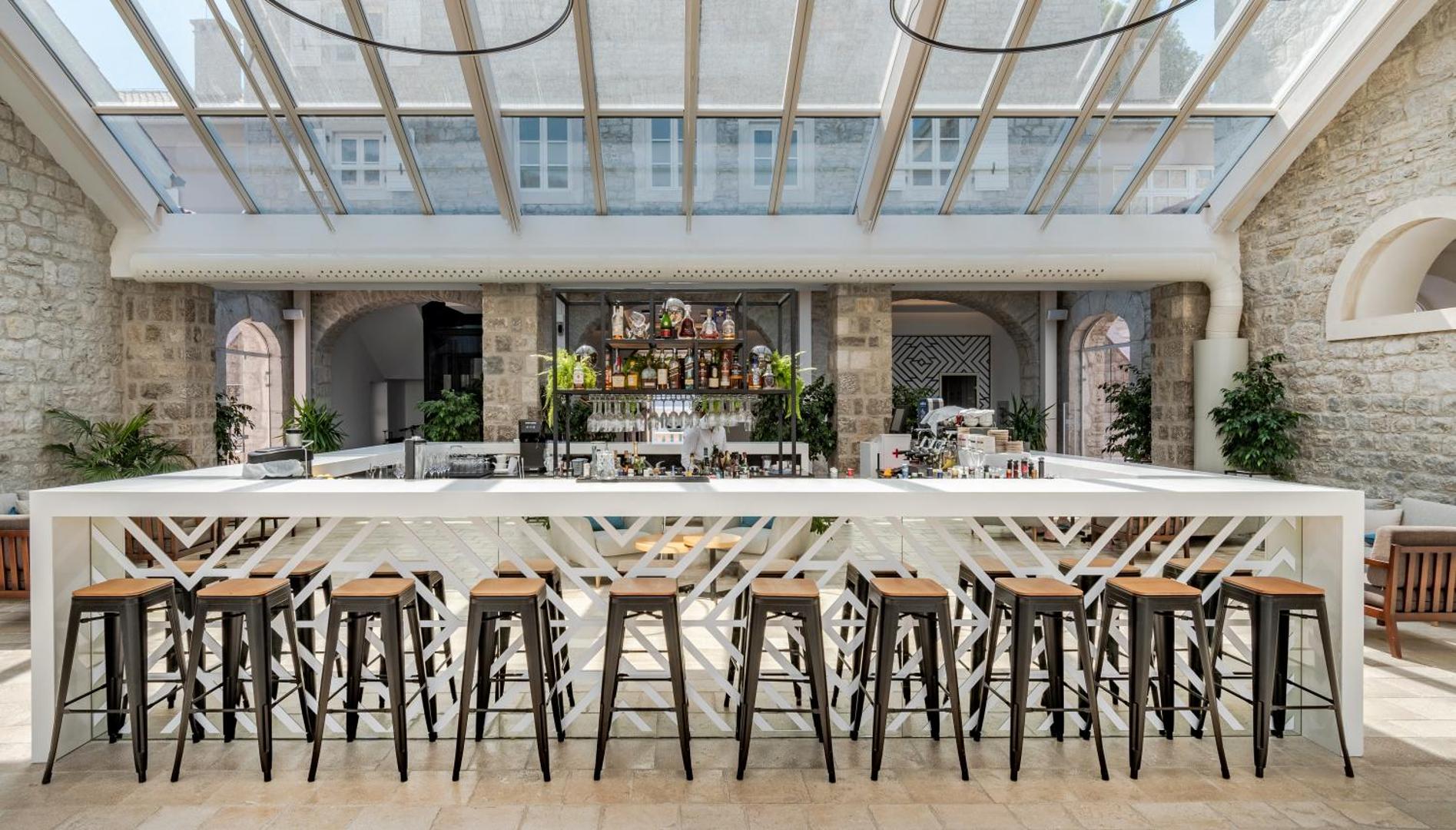 Lazure Hotel & Marina bārs ar modernu baltu leti un augstiem krēsliem zem stiklota jumta vēsturiskā akmens ēkā Melnkalnē