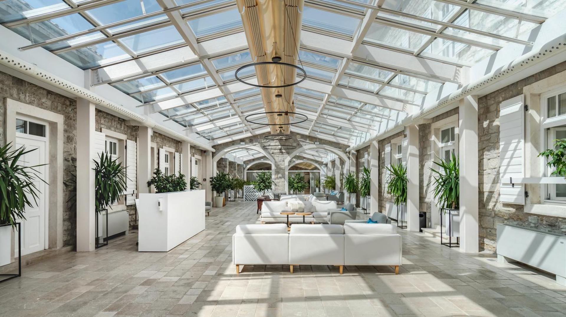 Lazure Hotel & Marina gaišais vestibils ar stikla jumtu, baltiem dīvāniem, akmens sienām un telpaugiem modernā dizainā