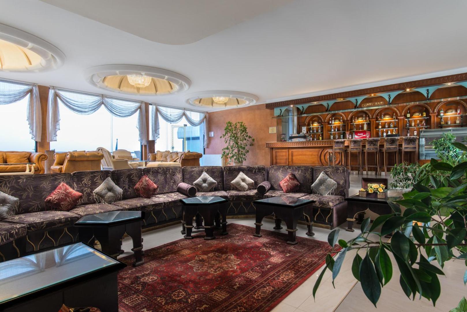 The Palace Hotel lobby bārs ar koka apdari, ērtiem dīvāniem un austrumu stila paklāju — mājīga atpūtas vieta viesnīcas iekštelpās