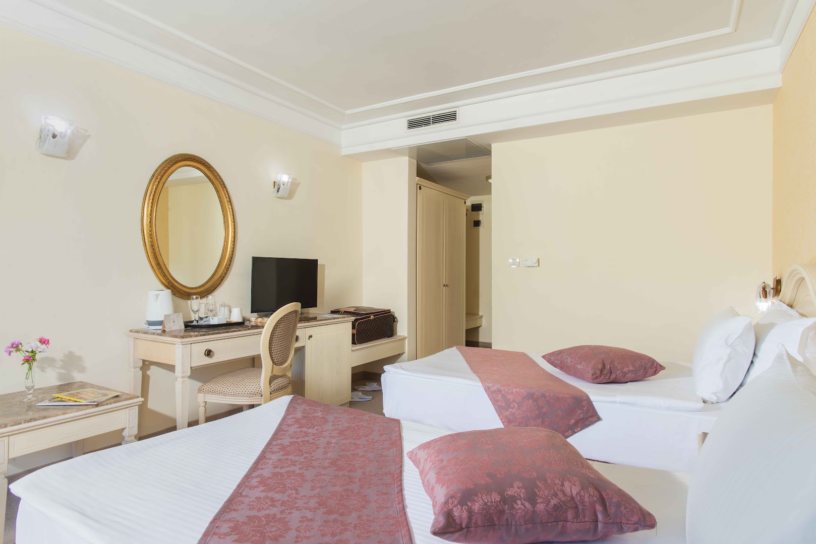 The Palace Hotel numura interjers ar divām gultām, rakstāmgaldu un spoguli — gaišas sienas un klasiska stila mēbeles
