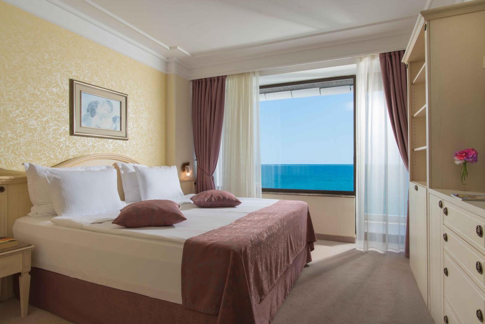 The Palace Hotel gaišs viesnīcas numurs ar divguļamo gultu, klasisku interjeru un lielu logu, caur kuru redzama jūra