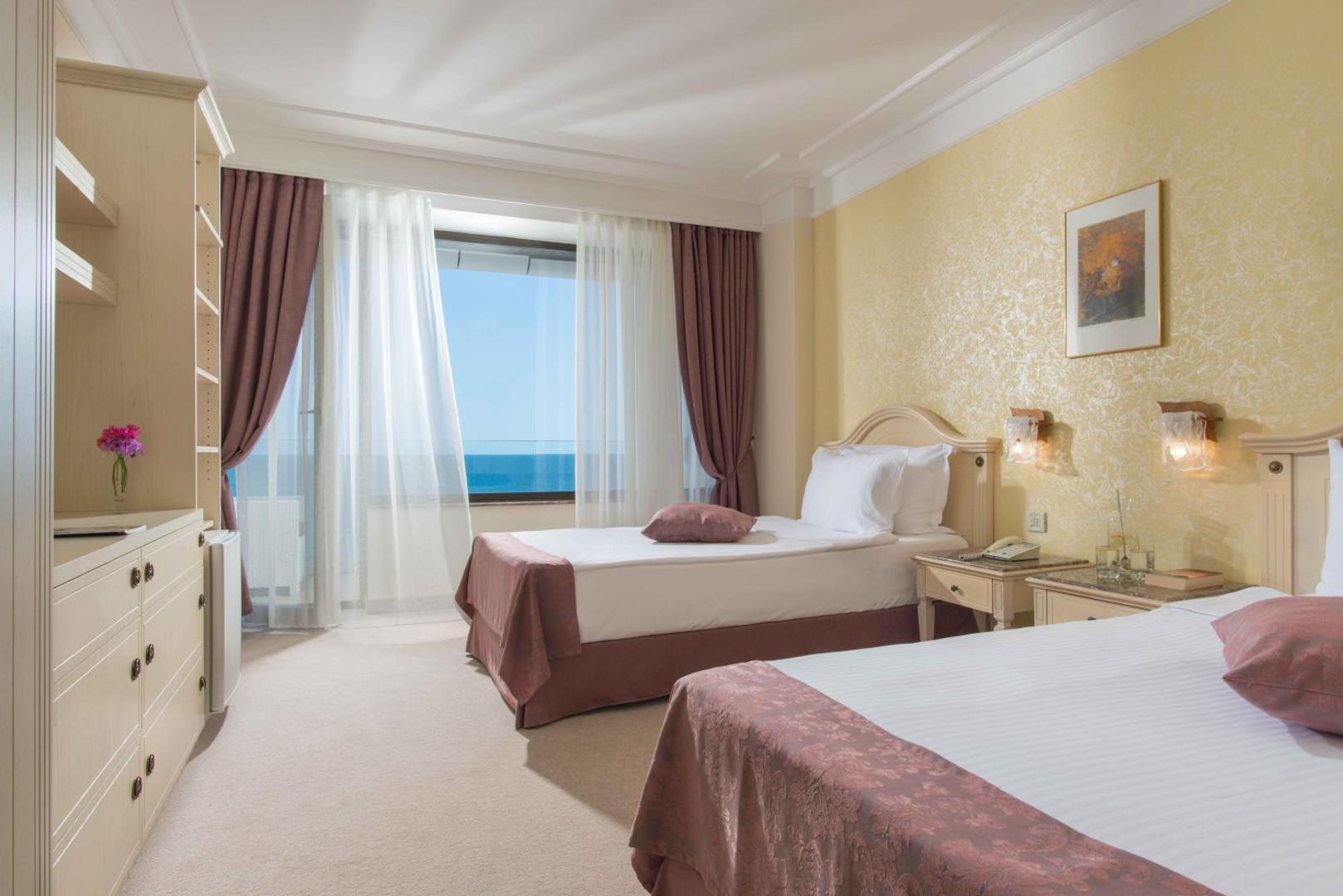The Palace Hotel gaišs numurs ar divām gultām un skatu uz jūru — klasisks interjers ar rozā akcentiem un lielu logu