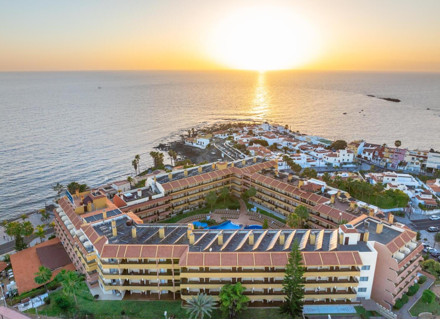 Hovima Jardin Caleta viesnīcas pagalms ar baseinu un saulessargiem no augšas — daudzstāvu numuru korpusi Tenerifes piekrastē saulrieta gaismā