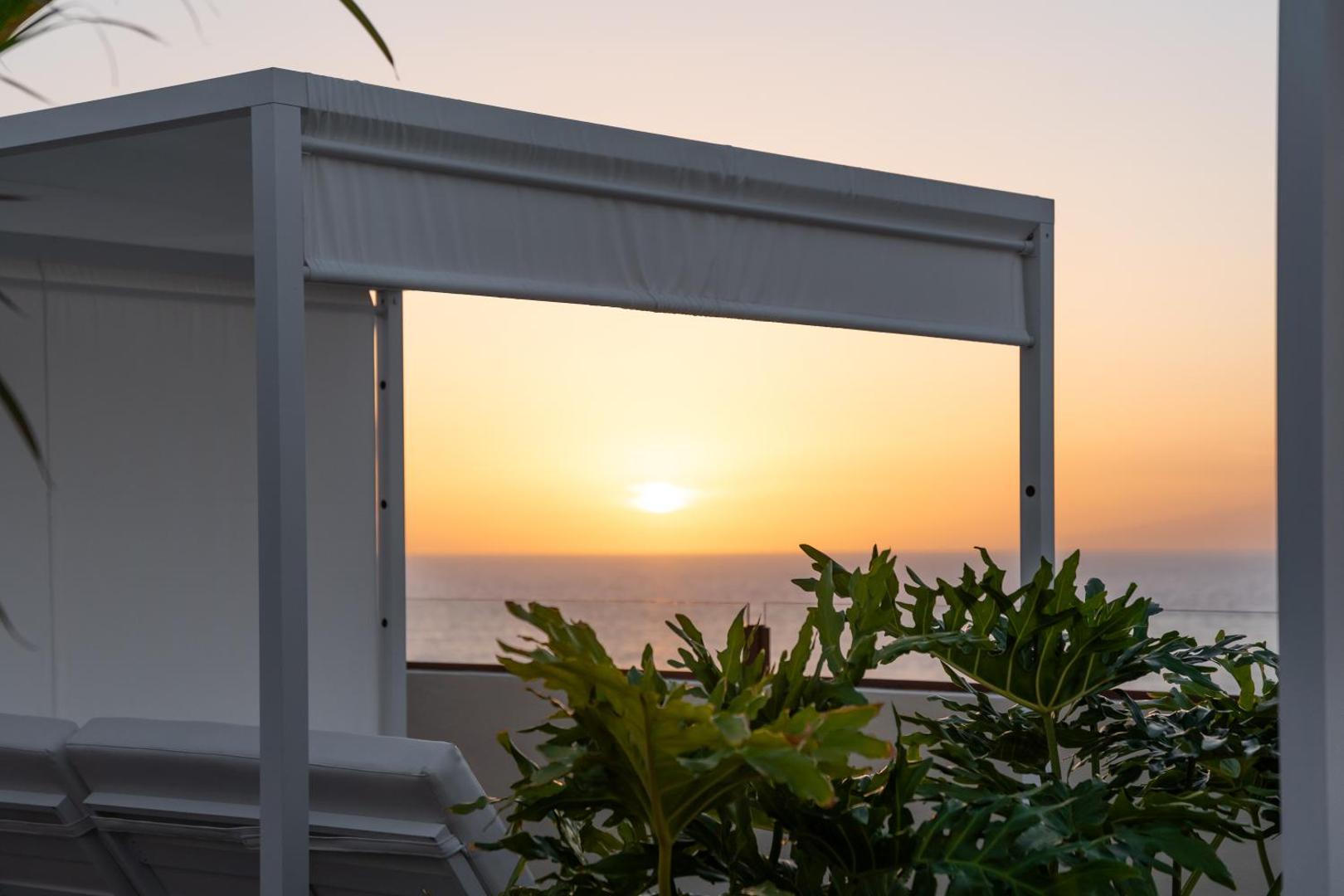 Skats uz saulrietu virs okeāna no Hovima Jardin Caleta terases Tenerifē caur baltu atpūtas nojumi un tropiskiem augiem