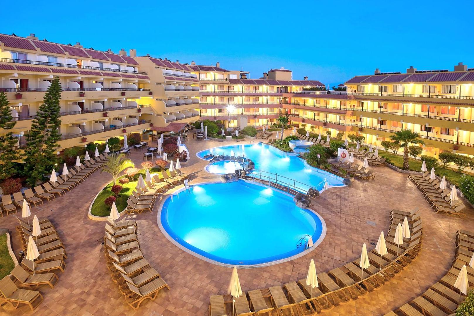 Hovima Jardin Caleta viesnīcas pagalms vakarā ar izgaismotiem baseiniem, sauļošanās krēsliem un daudzstāvu korpusiem Tenerifē.