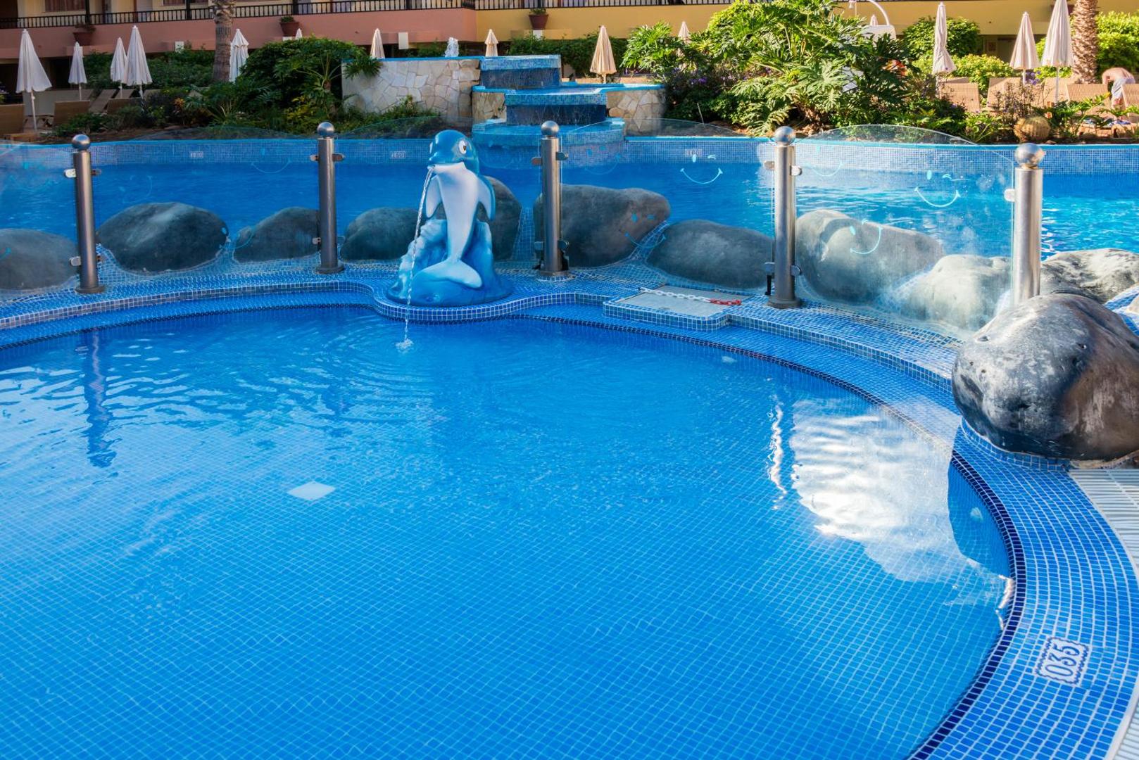 Hovima Jardin Caleta bērnu baseins Tenerifē ar zilu mozaīkas apdari, dekoratīvu delfīna strūklaku un drošības nožogojumu ģimenes atpūtai