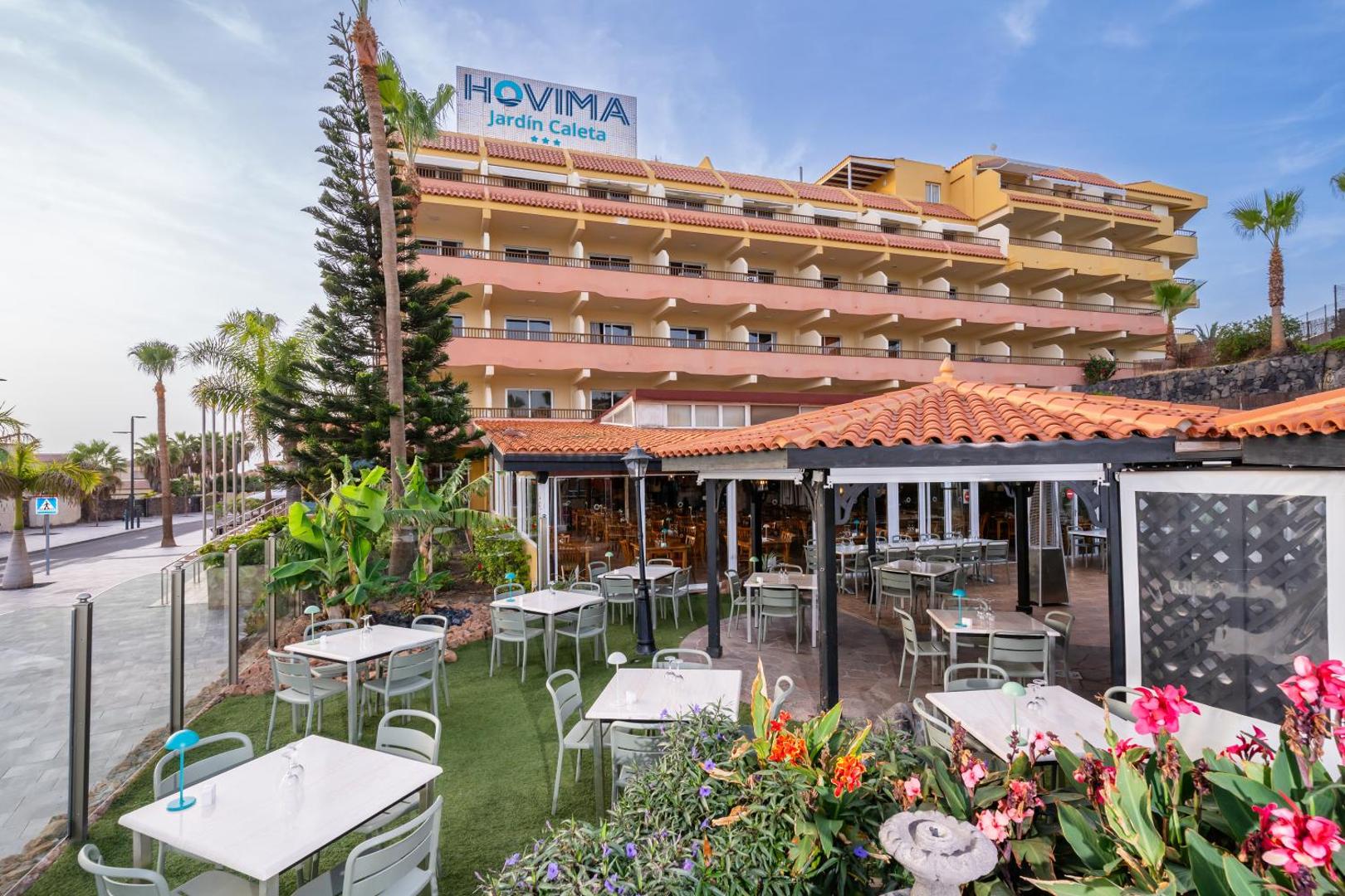 Hovima Jardin Caleta viesnīcas ēka Tenerifē ar āra restorāna terasi, galdiņiem uz zaļas zonas un ziedošiem augiem