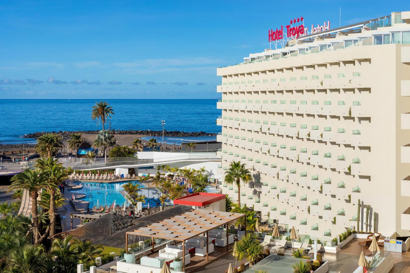 Alexandre Hotel Troya skats no augšas uz baseinu, terasi un okeāna piekrasti Tenerifē — viesnīcas ēka un atpūtas zona ar palmām