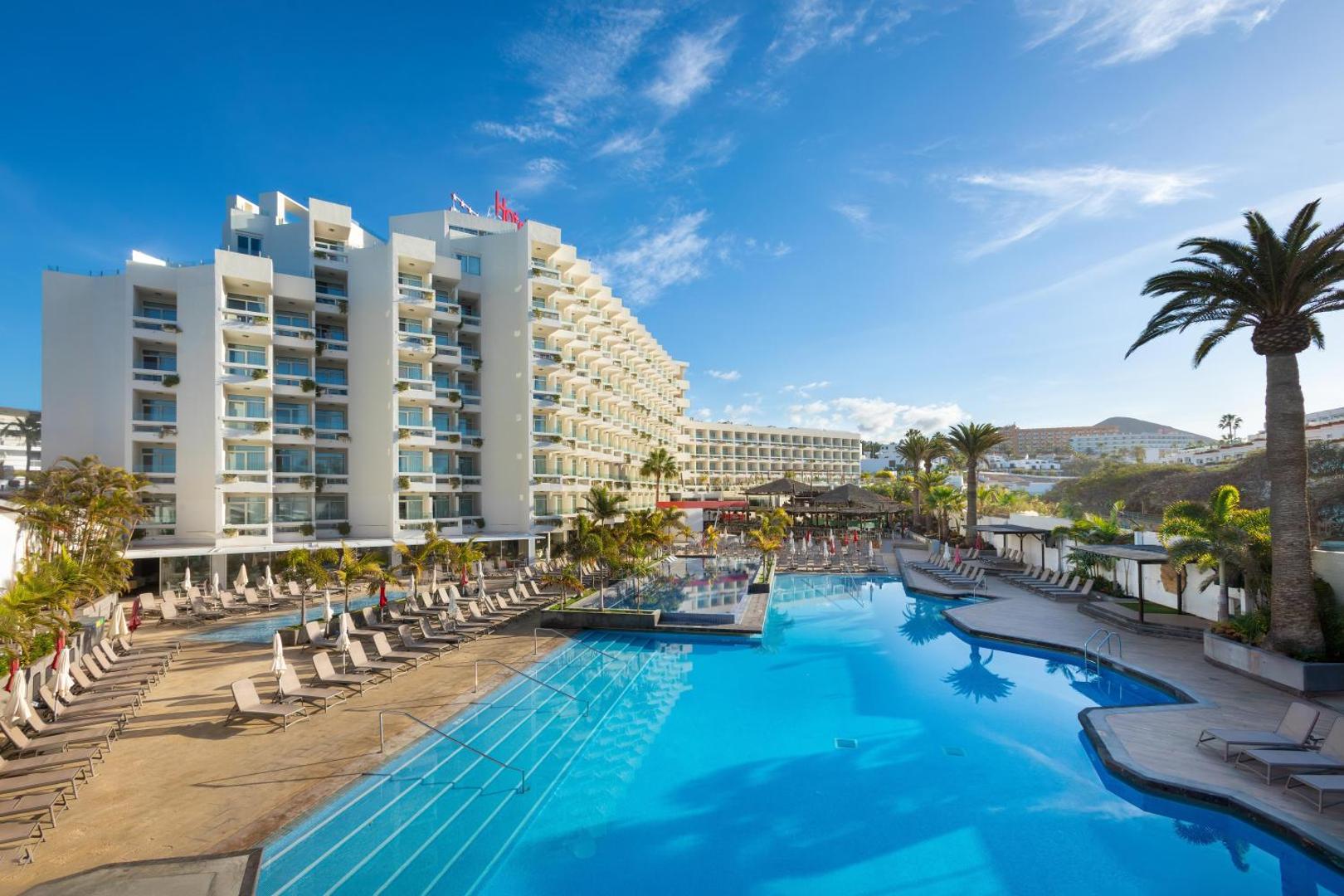 Alexandre Hotel Troya viesnīcas ēka un plašais āra baseins Tenerifē dienas laikā — apkārtnē palmas, sauļošanās krēsli un saulains debess fons.